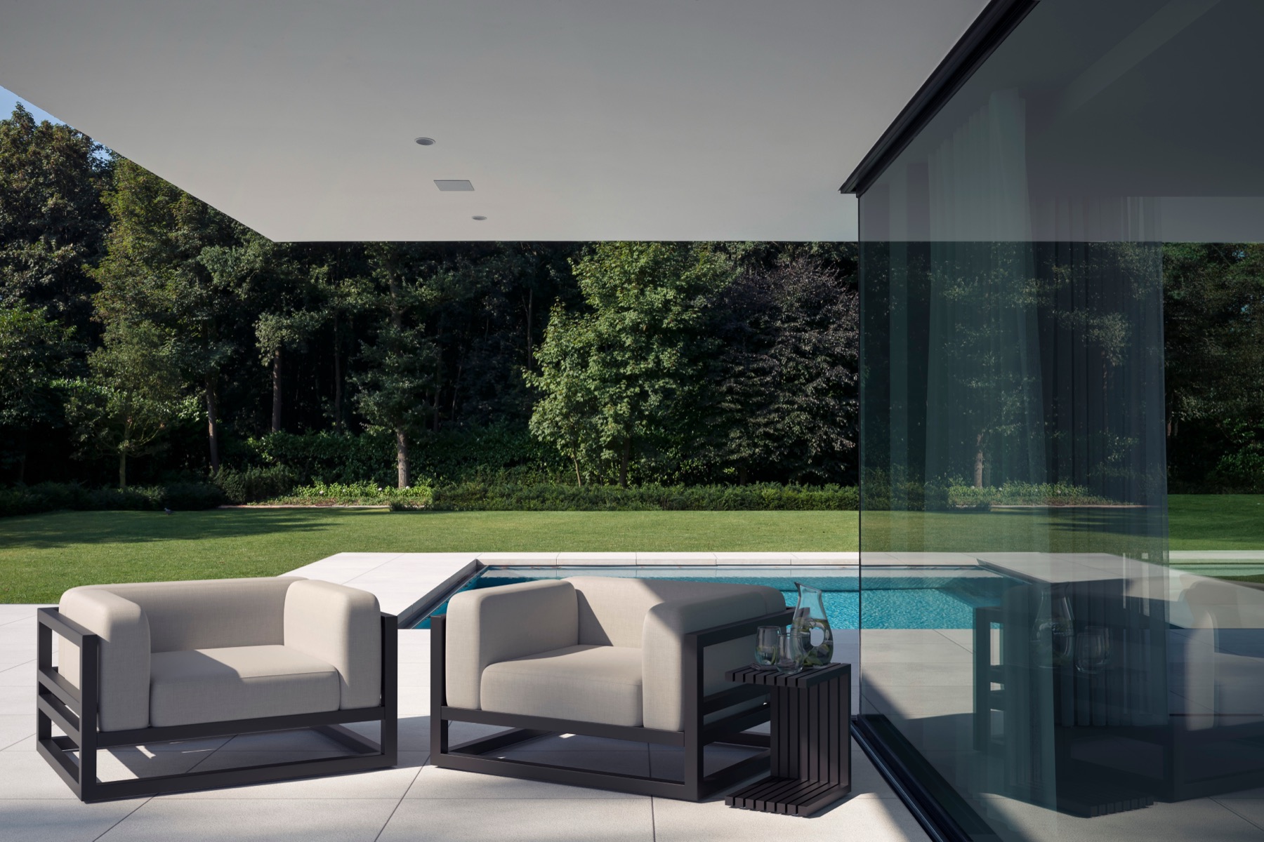 Two upholstered outdoor armchairs on a stone terrace beside a pool, sheltered by a cantilevered roof and looking out to a garden.